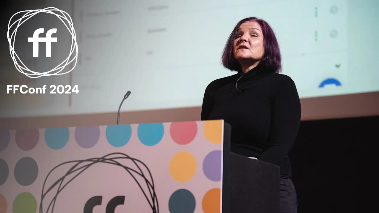 A woman, Léonie, with chin-length, vibrant purple hair and a black mock-neck top. She is standing behind a lectern, looking slightly upward and speaking.