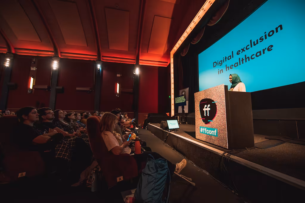 Sareh stood on stage in bright colours addressing the audience, with the screen behind her reading "Digital exclusion in healthcare".