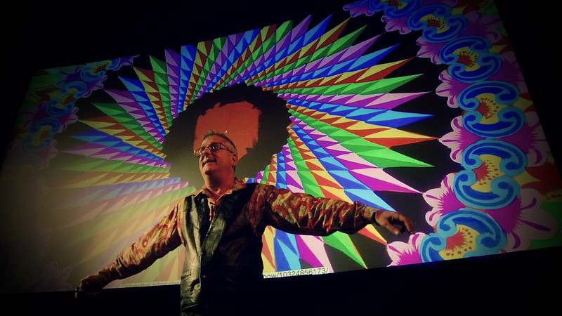Bruce Lawson stands in front of the large screen flowering out a psychedelic pattern like a tripping out peacock. He wears a black waistcoat and equally trippy shirt with his arms aloft owning the stage.