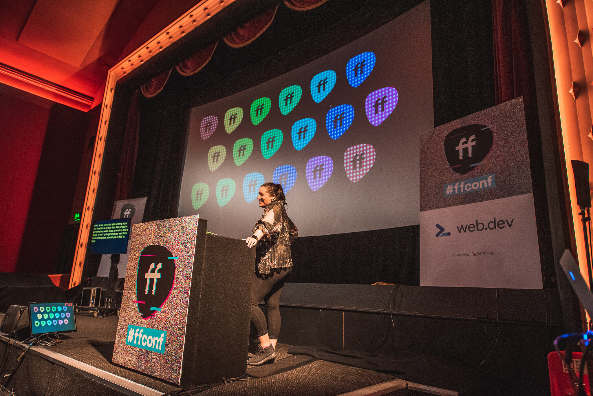 Ruth John stands behind a podium decorated with a static-textured overlay and the red and blue "#ffconf" logo. She is wearing a dark patterned top and dark trousers, facing towards the audience inside a classic theatre stage with warm lighting. Behind her, a large presentation screen displays rows of brightly coloured guitar pick shapes in shades of pink, green, blue, and purple, each containing the letters "ff."