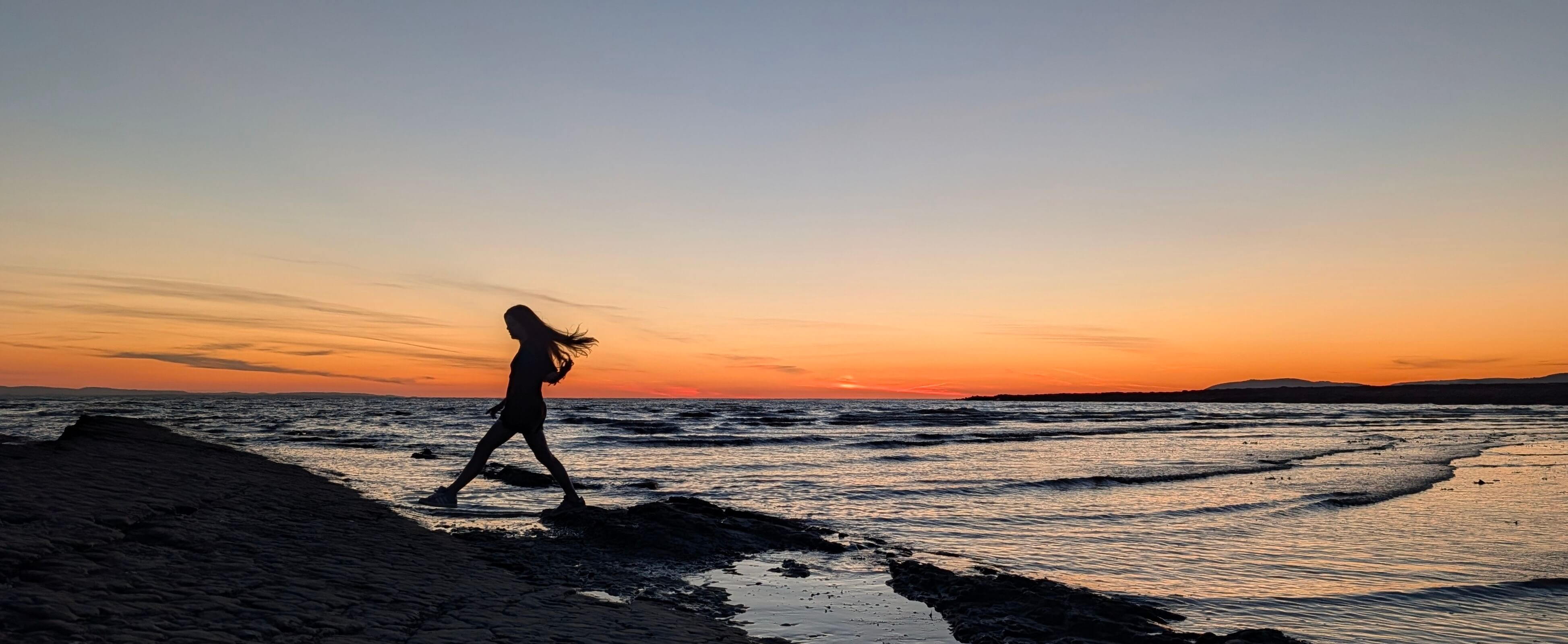 Silhouette of a long-haired person stepping across rocky tidal flats along a coastline under a vivid orange sunset sky